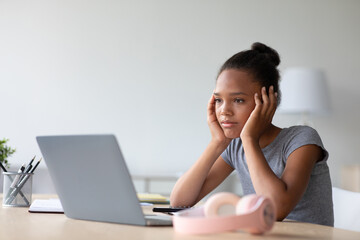 A pretty African American teenage girl sits at a desk in her room, wearing headphones while studying on a laptop. She looks sad and tired, showing the stress of remote education during the pandemic.