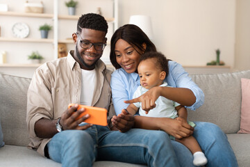 Young African American parents sit on a sofa at home, smiling as they show a funny educational app on a cellphone to their small son. The mother holds the baby, pointing at the screen.