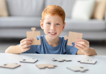 A joyful redhead boy holds two wooden puzzle pieces, smiling brightly. He engages with educational games at home, enjoying his time with smart activities designed for kids.