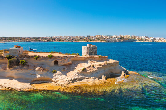 St. Thomas Bay in Malta, historic stone watchtower atop rocky headland overlooking turquoise sea and distant town. Natural beauty and historical significance of Mediterranean location