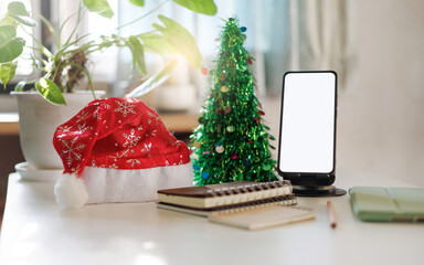 working on christmas holidays concept: close up view of mobile phone, red hat, notebook and christmas tree decoration on table
