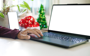 close up of man hand using laptop working with mobile phone and decoration christmas background with red hat, christmas tree on desk, working on christmas holiday concept