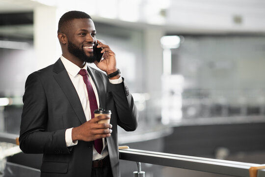 Cheerful young black bearded man manager standing at office building corridor, drinking coffee to go, having phone conversation with business partner and smiling, panorama with copy space