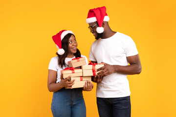 An African American couple smiles at each other while holding wrapped Christmas gift boxes. They wear red Santa hats, celebrating the festive spirit over a bright yellow background.
