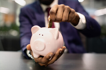 Savings, investment, business profit concept. Black businessman putting money in pink piggy bank, office interior, cropped. Unrecognizable african american man in suit making savings, closeup