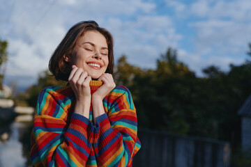 Young woman early 20s with short hair in a colorful striped sweater smiling with eyes closed, hugging collar outdoors by wooden railing in nature. Cozy joyful travel lifestyle.