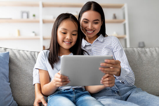 A joyful Asian mother and her young daughter sit on a comfortable couch at home, happily exploring a digital tablet together. - Powered by Adobe