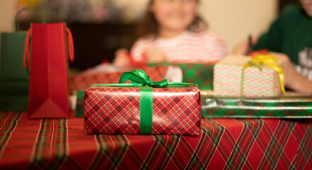 A festive scene with colorful wrapped box, holiday shopping bags, and a cozy plaid tablecloth, evoking the joy of Christmas gift-giving. In the background children are decorating gifts.