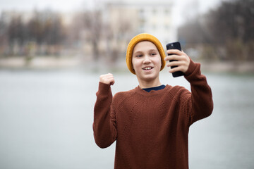A cheerful teenager kid boy takes a selfie with a smartphone or recording video blog outdoors near a lake or pond, urban landscape in the background.
