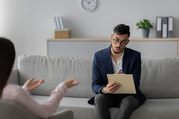 An Arab male psychologist conducts a psychotherapy session with a young woman. He attentively listens and takes notes while she shares her thoughts in a cozy office environment.