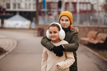 Two children siblings hugging outdoors in winter, wearing warm clothes, smiling happily in a city park.