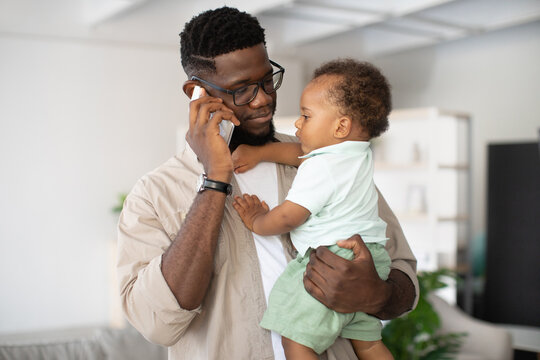 A black father smiles as he talks on his cellphone while holding his toddler in his arms. He is balancing work responsibilities and spending quality time at home with his son.
