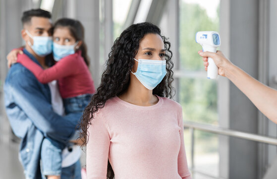 A family stands in a public area while a worker checks the temperature of an adult woman. All individuals are wearing face masks for safety.