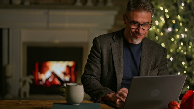 Happy smiling mid adult man using laptop computer at home, sitting at desk in Christmas decorated cosy room. Fireplace and Christmas tree in background. Businessman shopping online searching for gift.