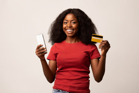 A cheerful African American woman smiles while holding her smartphone and credit card, using an application for online shopping.