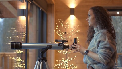 Middle aged woman at home looking out window with telescope. Female in turtleneck pullover standing in living room of wooden cabin with Christmas lights. Autumn or winter indoor clothing.