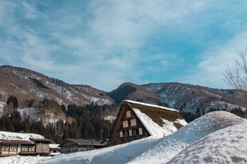 青空と雪山を背景にした合掌造りの家屋 © tokoteku_2018