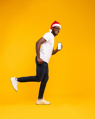 A cheerful black man wearing a Santa hat shows a blank phone screen. He stands against a vibrant yellow background. The scene captures the holiday spirit and the excitement of mobile offers.