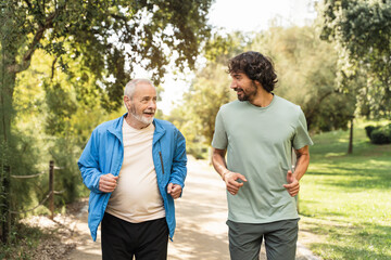 Healthy senior man practicing sport, together with young adult exercising together, enjoying conversation while running in a park
