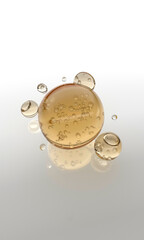 A realistic 3D rendered image featuring several golden or amber-colored oil serum drops (or capsules) suspended in the air and resting on a white, reflective surface.