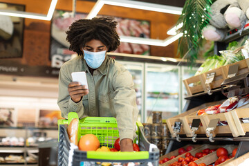 A young man wearing a mask selects fresh fruits and vegetables in a grocery store. He checks his phone for a shopping list while filling his cart with healthy food choices.