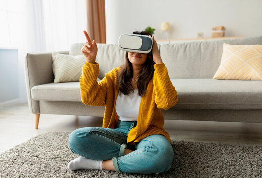 Joyful arab woman wearing VR glasses sits on the floor of her living room, pointing at unseen elements. She is engaged in a virtual reality session, enjoying her leisure time at home.