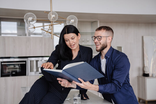 A couple enthusiastically engages with a large book in a modern kitchen, sharing a moment of curiosity and connection.