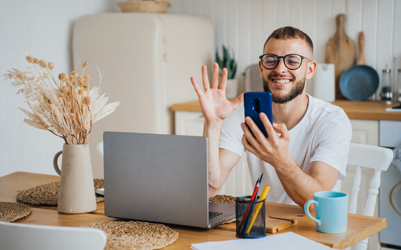 A smiling man waves at his phone from the dining table, laptop open, exuding a warm morning energy in a homey kitchen setting.