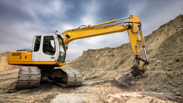 A yellow excavator operates on a construction site, digging and moving earth. Dust rises as it works on the rugged terrain under a cloudy sky