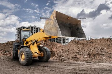 A large bulldozer loader lifts a pile of dirt on a construction site surrounded by buildings and...