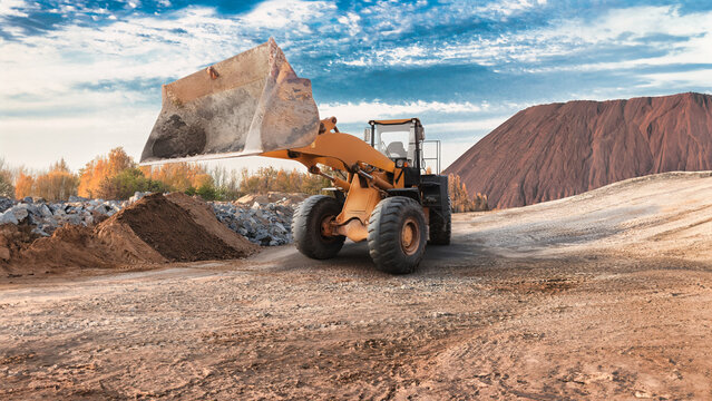 A large wheel loader removes soil from a site surrounded by hills. The sky is clear, and the trees are painted in autumn colors, heralding the arrival of autumn