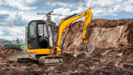 Mini excavator is actively digging at a busy construction site, with towering buildings and a crane...