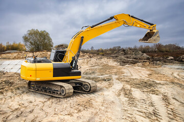A yellow excavator is working at a construction site in an urban area, moving dirt and preparing the ground under overcast skies