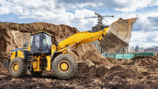 A large yellow wheeled bulldozer or loader is digging and relocating earth at a busy construction site surrounded by buildings and cranes under a dramatic sky