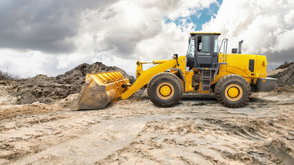 Heavy machinery moves dirt at a construction site. A yellow loader uses its bucket to lift soil. Fluffy clouds fill the sky, creating an overcast afternoon scene