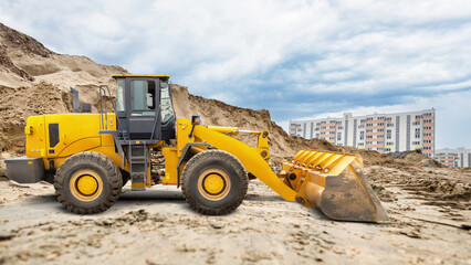 A yellow loader works at a construction site, scooping sand with its bucket near newly built residential apartments on a cloudy day