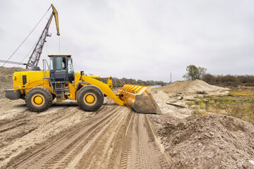 Obraz premium A large yellow loader moves sand at a construction area beside a river. The ground shows tire tracks, hinting at active work. The sky is overcast