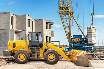 Yellow loader and crane operate on a construction site, moving heavy materials among partially built structures under a clear sky