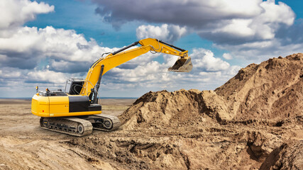 Obraz premium An excavator operates on a construction site, digging into a large mound of dirt under a bright blue sky with fluffy clouds in the background