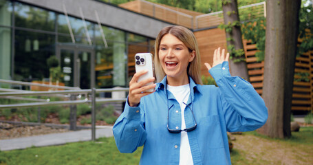 Happy blonde woman walking in park and talking online with friends using smartphone in front of modern glass building. Communication and lifestyle concept
