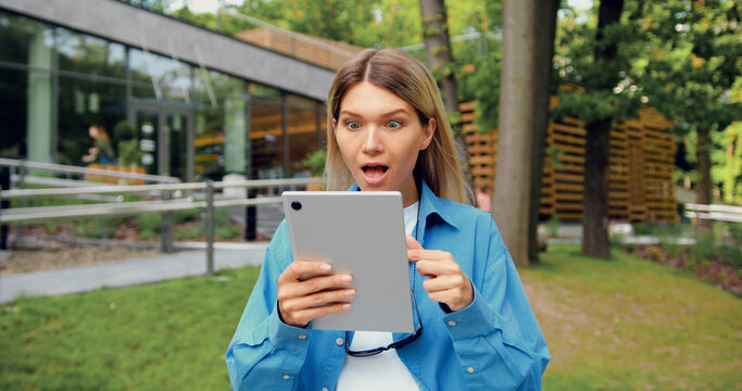 Surprised Caucasian woman using digital tablet outdoors in urban park, reacting to sudden news while looking at screen. Emotions and communication concept. - Powered by Adobe