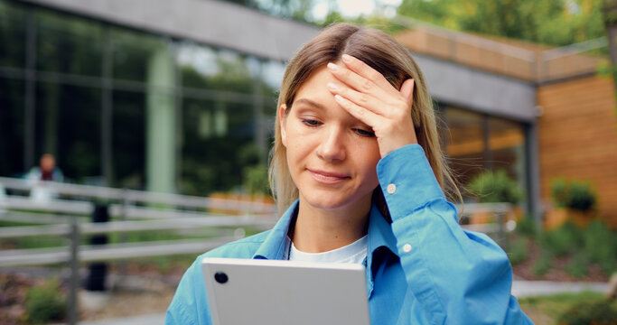 Portrait of anxious female holding tablet in hands and looking worried at screen outdoors in urban park. Concern and communication concept.