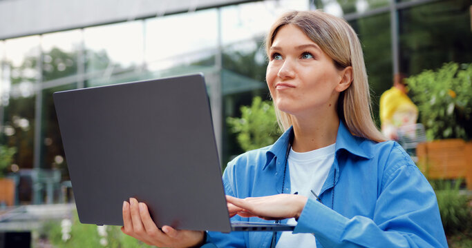 Smiling professional businesswoman typing on a laptop outdoors near a modern office building. Remote work concept, digital communication, corporate lifestyle, urban business environment.