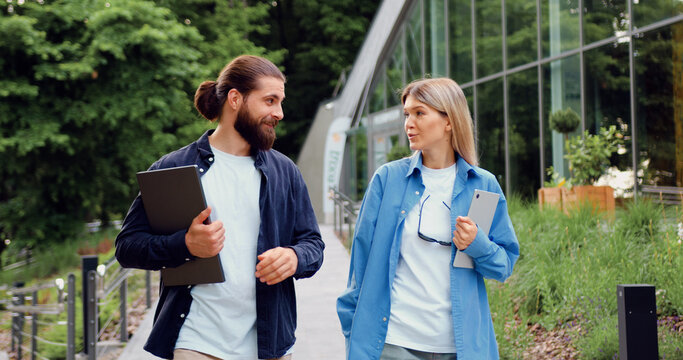 Two business professionals, man with laptop and woman with tablet, wearing casual office attire, walking outside, while engaged in a work-related conversation, showcasing teamwork