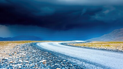 A gravel road curves through a barren, rocky landscape with sparse dry grass. Dark, dramatic storm clouds fill the sky, creating a moody and atmospheric scene.