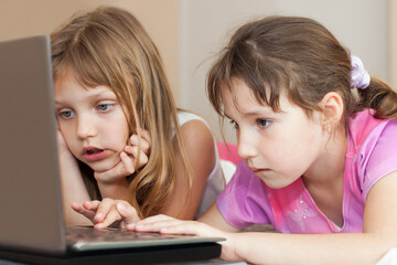 Close-up portrait of two young girls using a laptop indoors, focused and engaged while looking at the screen.