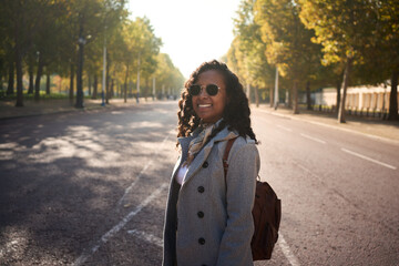 Cheerful African millennial in a gray coat and sunglasses smiles at the camera on a sunlit, tree-lined city street, with soft bokeh and autumn vibes suggesting travel and relaxed