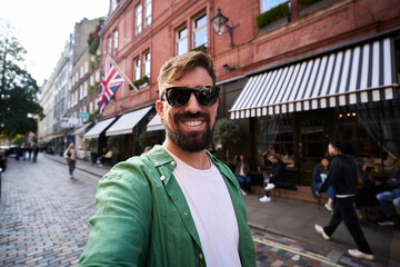 Smiling man taking a selfie looking at camera on a London street with a Union Jack and striped awnings behind him. Casual travel moment, daylight, urban vibes and cheerful tourism energy.