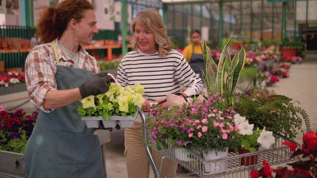 Young male employee assisting a senior female customer with her selection of potted plants at a large, bustling garden center, offering expert advice and friendly customer service - Powered by Adobe