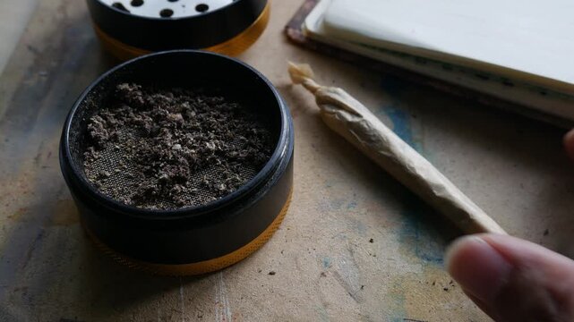 Hand holding rolled marijuana joint with grinder and cannabis herb on wooden table for recreational use
